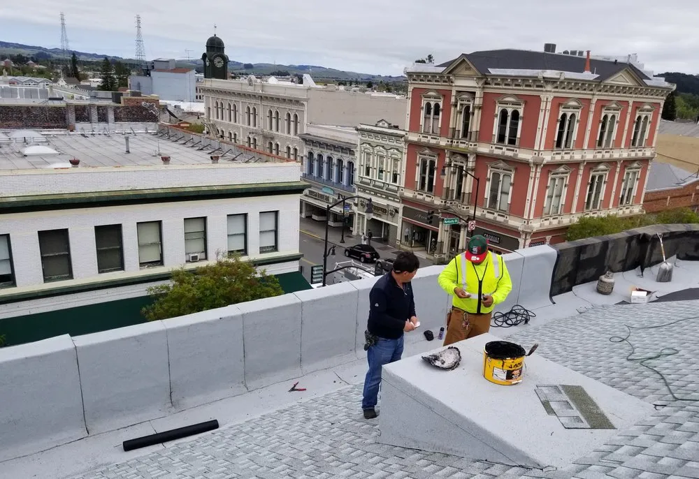 "Two workers from JK Pro Roofing, Inc. engaged in a rooftop repair in Daly City, surrounded by historical buildings and urban skyline."